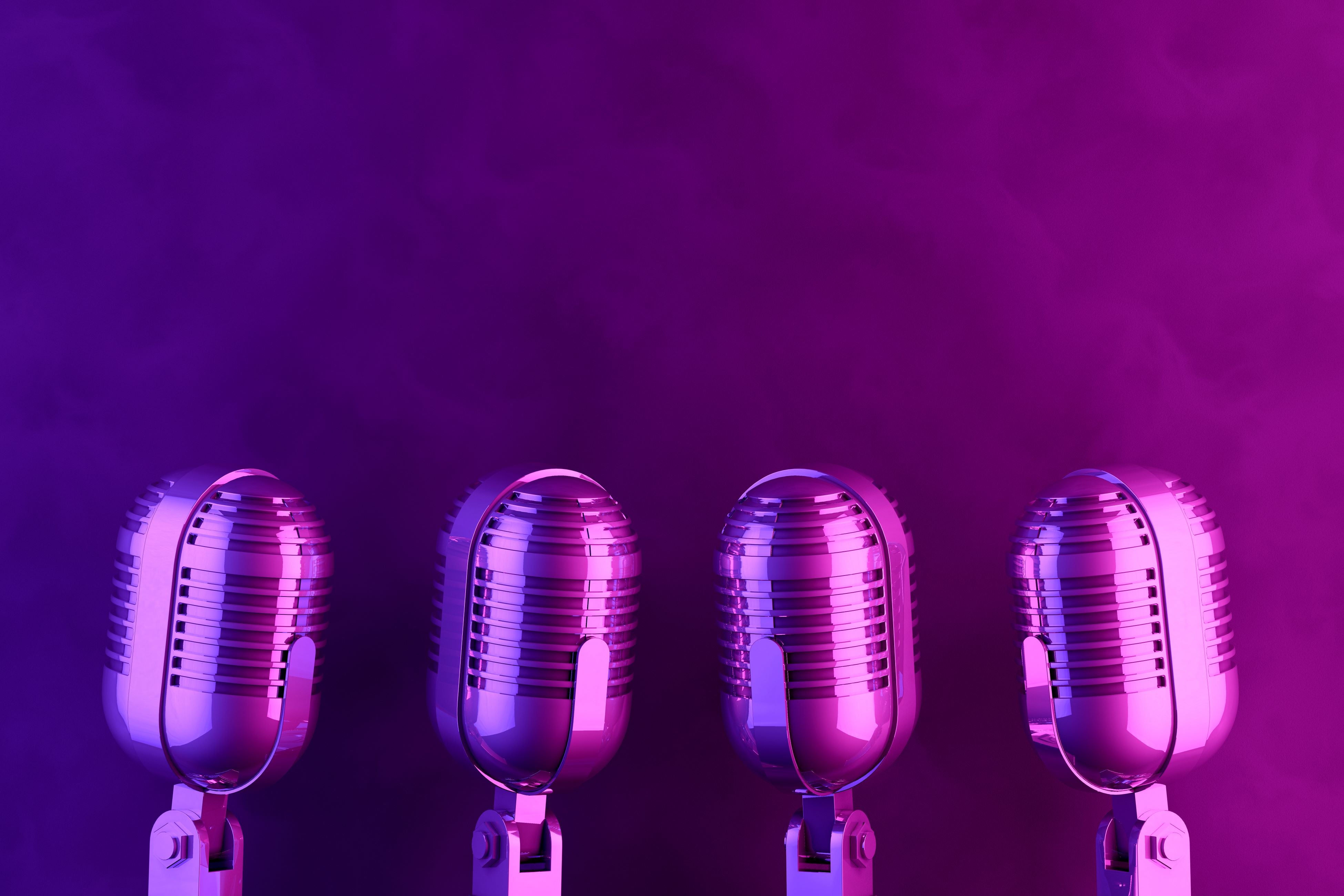 Four antique microphones reflecting purple light are lined up in a row in front of a purple background.