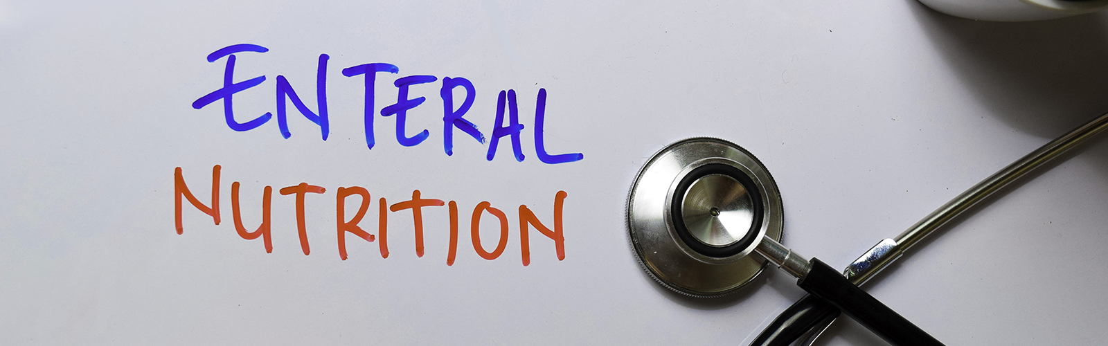 A silver stethoscope sits on top of a whiteboard with the words “Enteral Nutrition” written in blue and red marker ink. 
