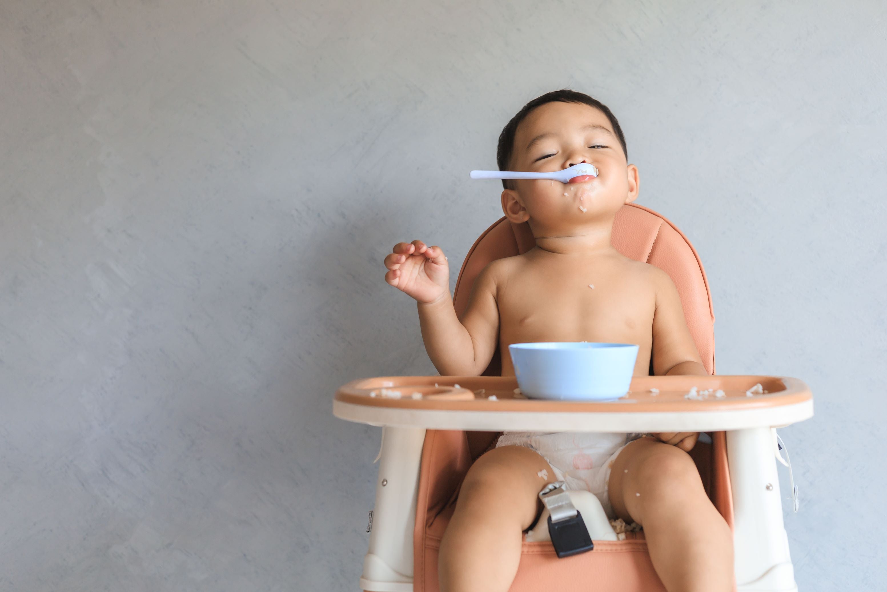 A baby seated in a highchair with a blue bowl on the tray smiles with a blue spoon in their mouth. 