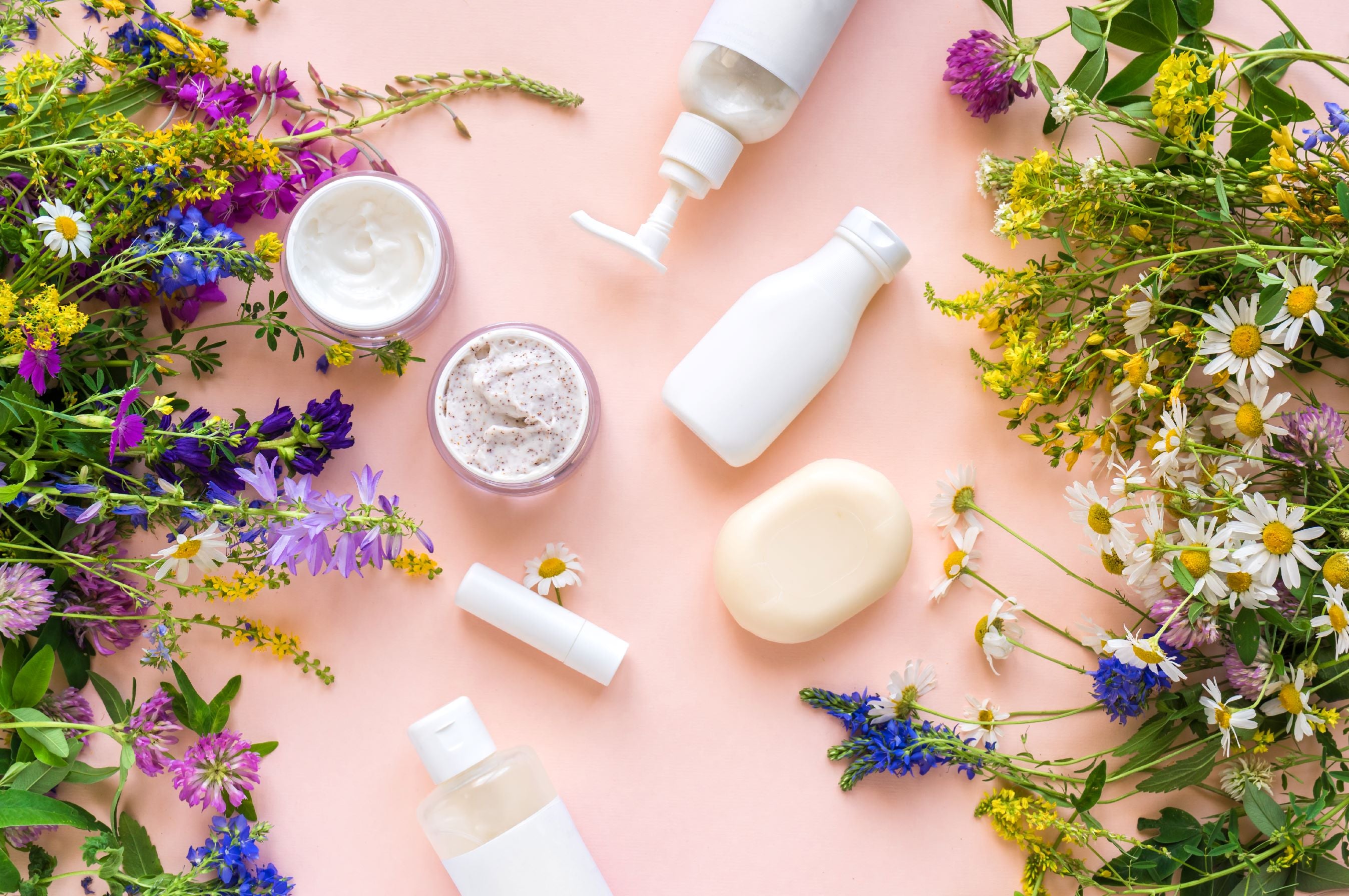 White bottles, a bar of soap and jars of lotion are displayed on a pink background with wildflowers.