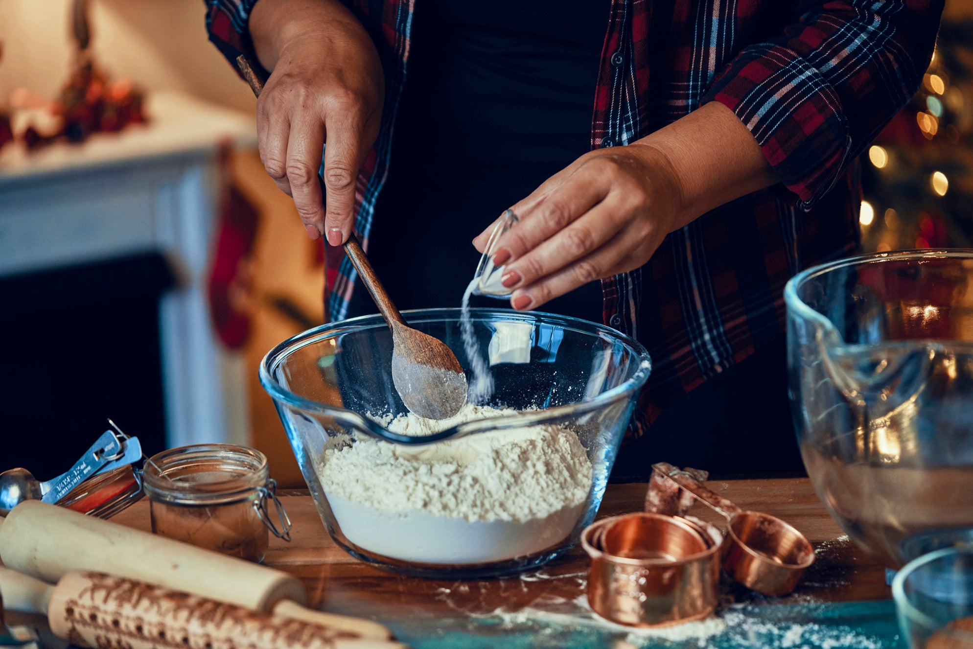 A person holding a wooden spoon pours baking soda into a bowl filled with flour on a table scattered with baking utensils such as measuring cups and rolling pins.