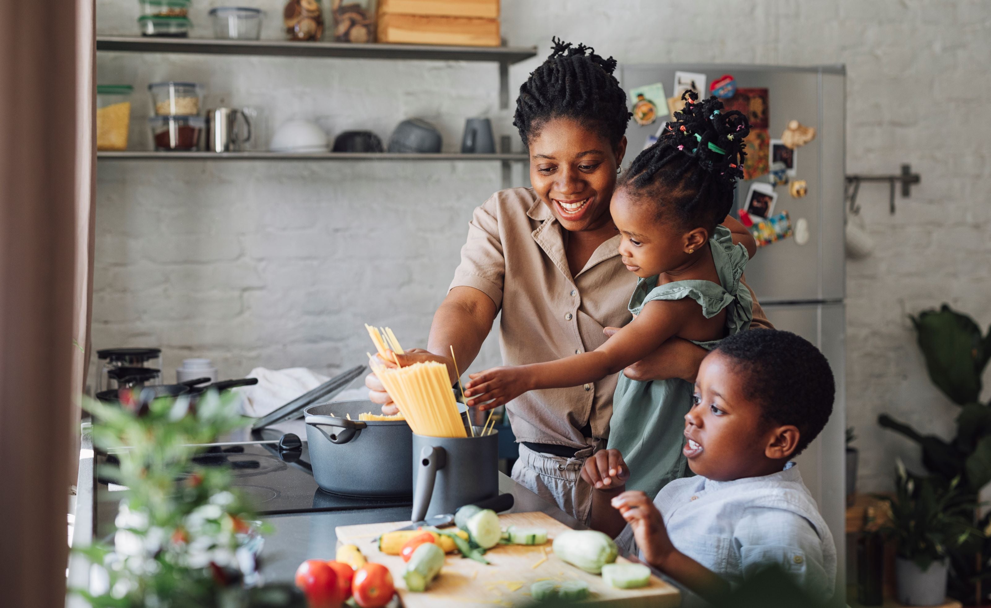 An adult and two children place dry spaghetti in a pot together in a kitchen.